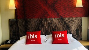 Quarto de hotel com uma cama grande, duas almofadas vermelhas com a inscrição "ibis HOTELS" e um painel de cabeceira com padrão marmorizado, além de duas luminárias pendentes.