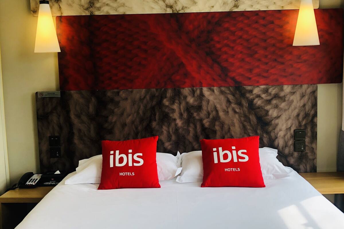Une chambre d’hôtel avec un grand lit, deux oreillers rouges ornés du texte « ibis HOTELS », une tête de lit à motifs de marbre et deux lampes suspendues.