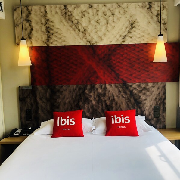Une chambre d’hôtel avec un grand lit, deux oreillers rouges ornés du texte « ibis HOTELS », une tête de lit à motifs de marbre et deux lampes suspendues.