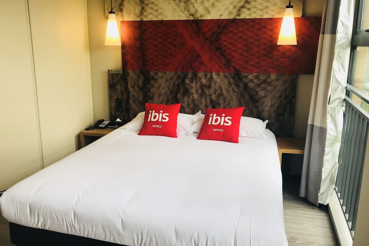 Een hotelkamer met een groot bed, twee rode kussens met de tekst "ibis HOTELS", een nachtkastje met een telefoon en een hoofdbord met een rood-bruin patroon.