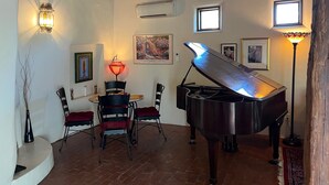 A cozy room with a grand piano, a dining table with chairs, a small window, and a wall-mounted air conditioner. - Balloon Fiesta close to Charming, Historic, N. Valley Casita! (Albuquerque)