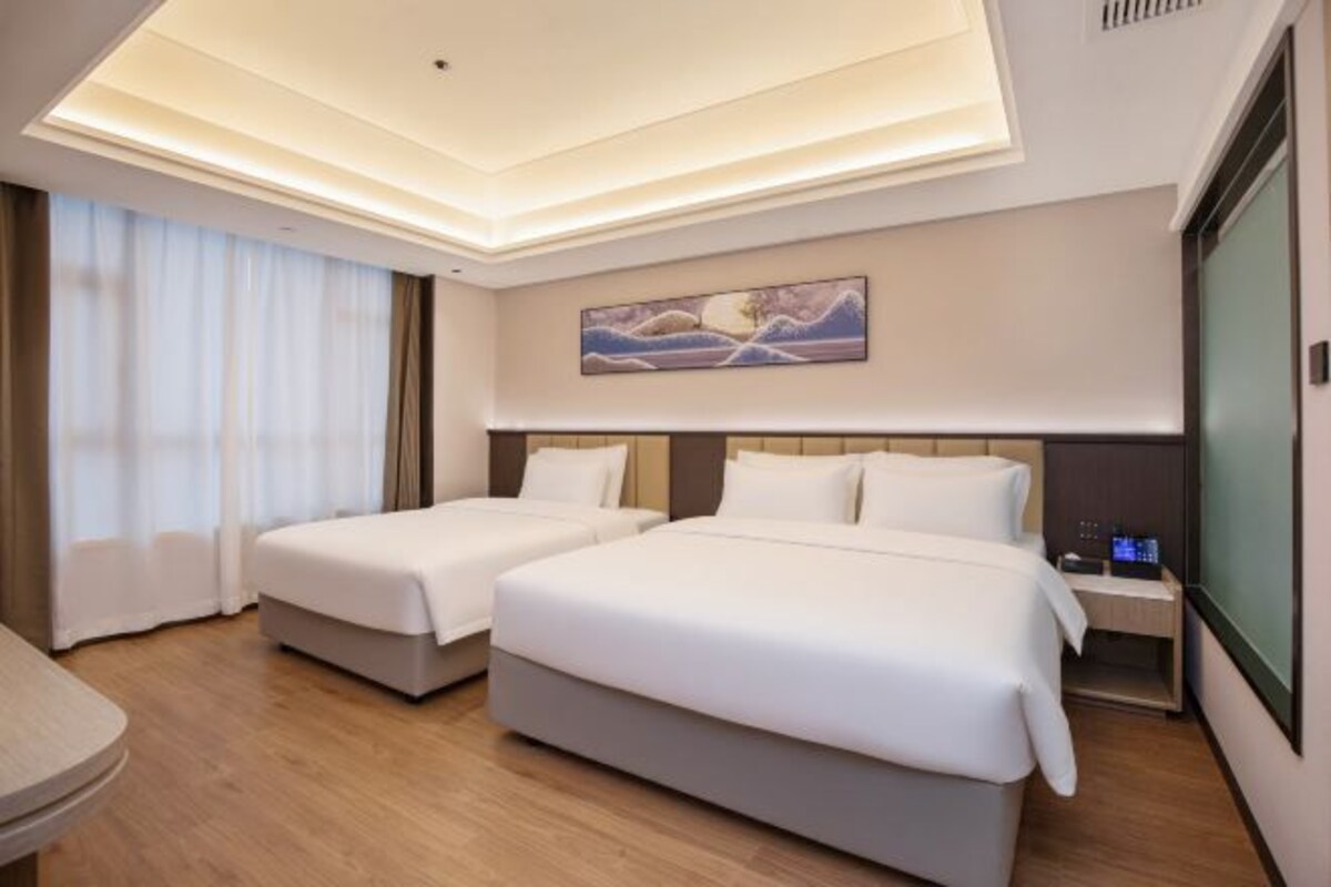 A hotel room with two beds, a painting on the wall, a desk with a lamp, and a window with curtains.