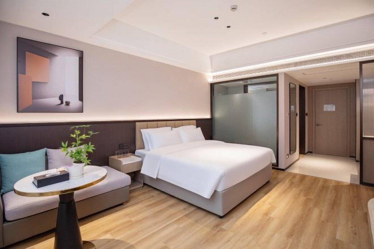 A modern hotel room with a large bed, a sofa, a small table with a plant, and a framed picture on the wall.