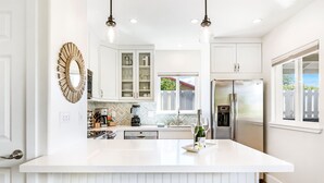 A modern kitchen with white cabinetry, a central island, pendant lighting, and a view of the outdoors. - Luxury cottage, private, saltwater spa🌺walk to beach, dining,shops (Kihei)