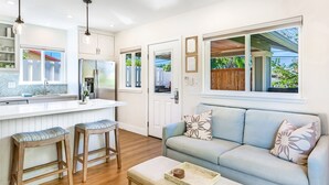 A modern kitchen with a white countertop, two bar stools, a wooden bench, a light blue sofa with floral cushions, and a wooden coffee table. - Luxury cottage, private, saltwater spa🌺walk to beach, dining,shops (Kihei)
