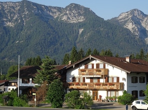 A traditional Alpine-style building with a balcony, surrounded by lush greenery and mountains in the background.