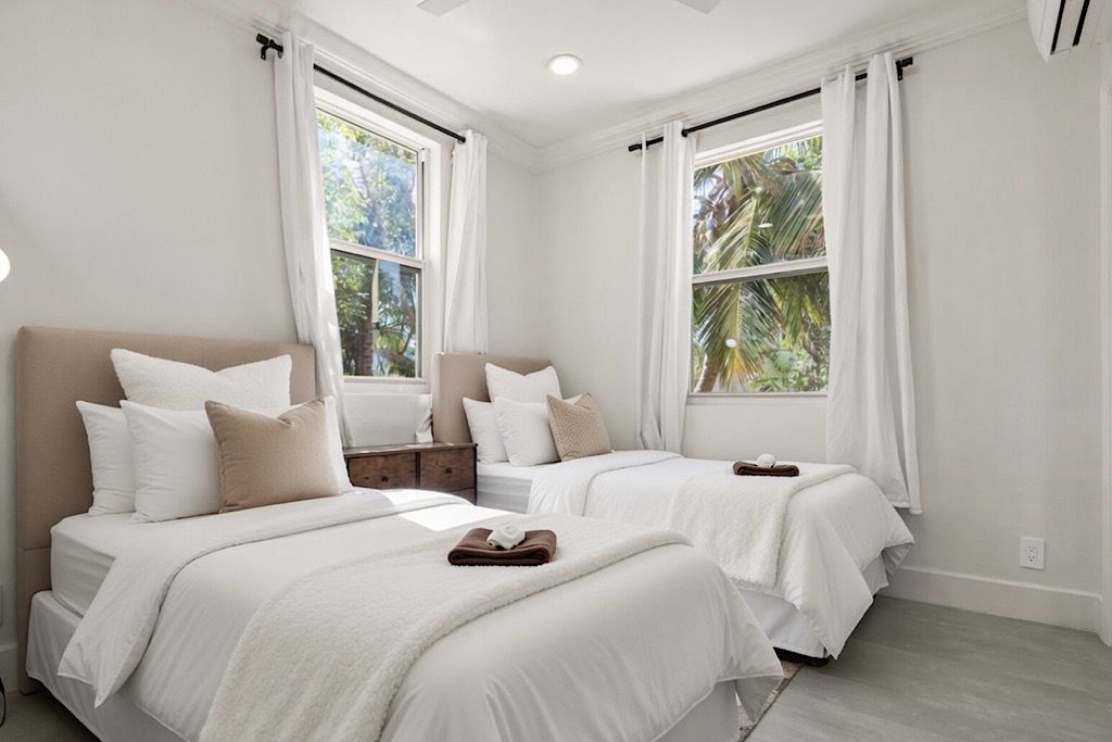 Habitación de hotel con dos camas, ropa de cama blanca, una mesita de noche de madera y ventanas amplias con cortinas.