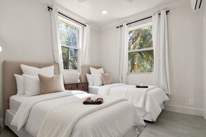 A hotel room with two beds, white bedding, a wooden nightstand, and large windows with curtains.