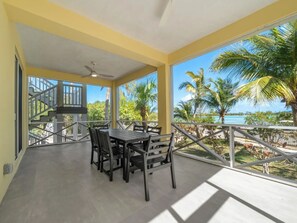 A covered balcony with a table and chairs, a ceiling fan, and a view of palm trees and a beach.