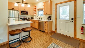 A compact kitchen with wooden cabinets, stainless steel appliances, and a white countertop. There are two stools and a small bar area. - Fisherman’s Cabin in Soldotna Near Kenai River! (Soldotna)
