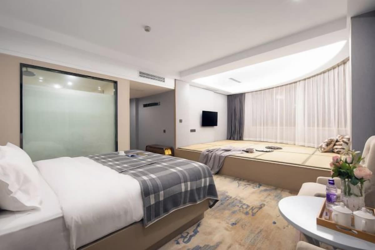 A modern hotel room with a bed, a seating area with a table, a flat-screen TV, and a large window with curtains.
