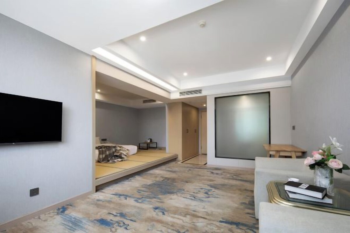 A modern hotel room with a flat-screen TV mounted on the wall, a bed with a headboard, a small bedside table, and a glass coffee table with a vase of flowers.