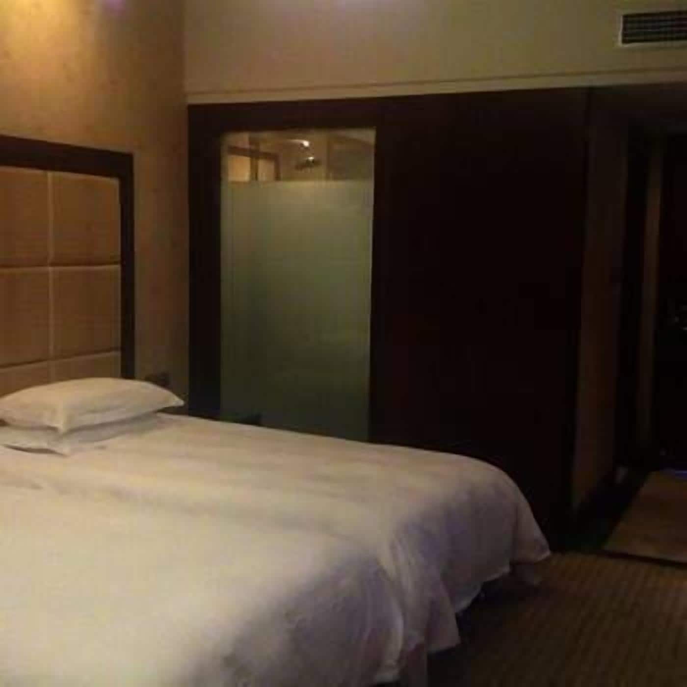 A hotel room with a bed, a wooden headboard, a glass shower enclosure, and a dark-colored door.