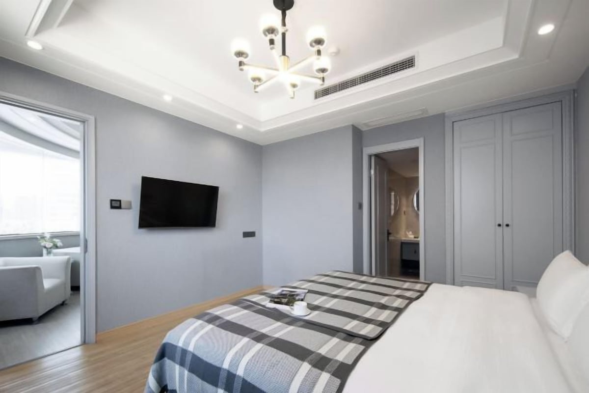 A modern bedroom with a large bed, a flat-screen TV mounted on the wall, and a ceiling light fixture.