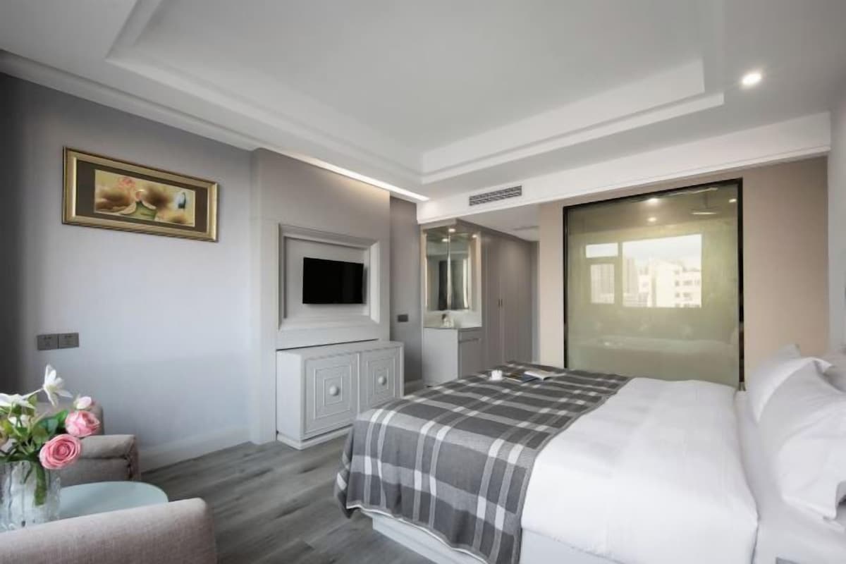 A modern bedroom with a large bed, a flat-screen TV mounted on the wall, a vanity with a mirror, and a window with a view of buildings.