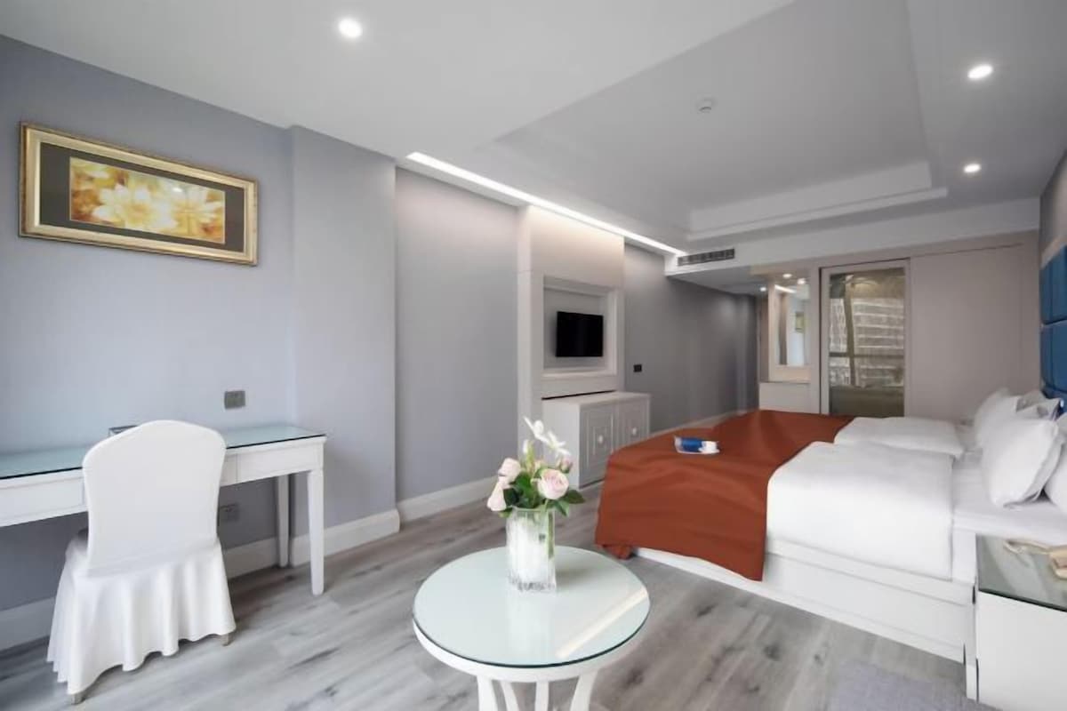 A modern hotel room with a bed, a desk, a chair, a glass table, and a vase with flowers.