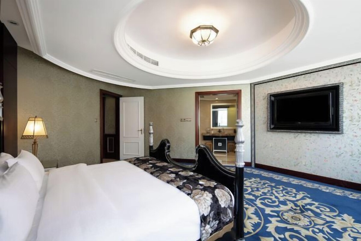 A hotel room with a large bed, a desk, a chair, a lamp, a television, and a patterned carpet.