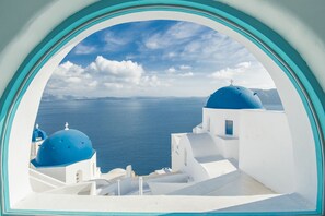 A view through a window of white buildings with blue domes, overlooking a body of water and distant land. - Sophia Oia View (Santorini)
