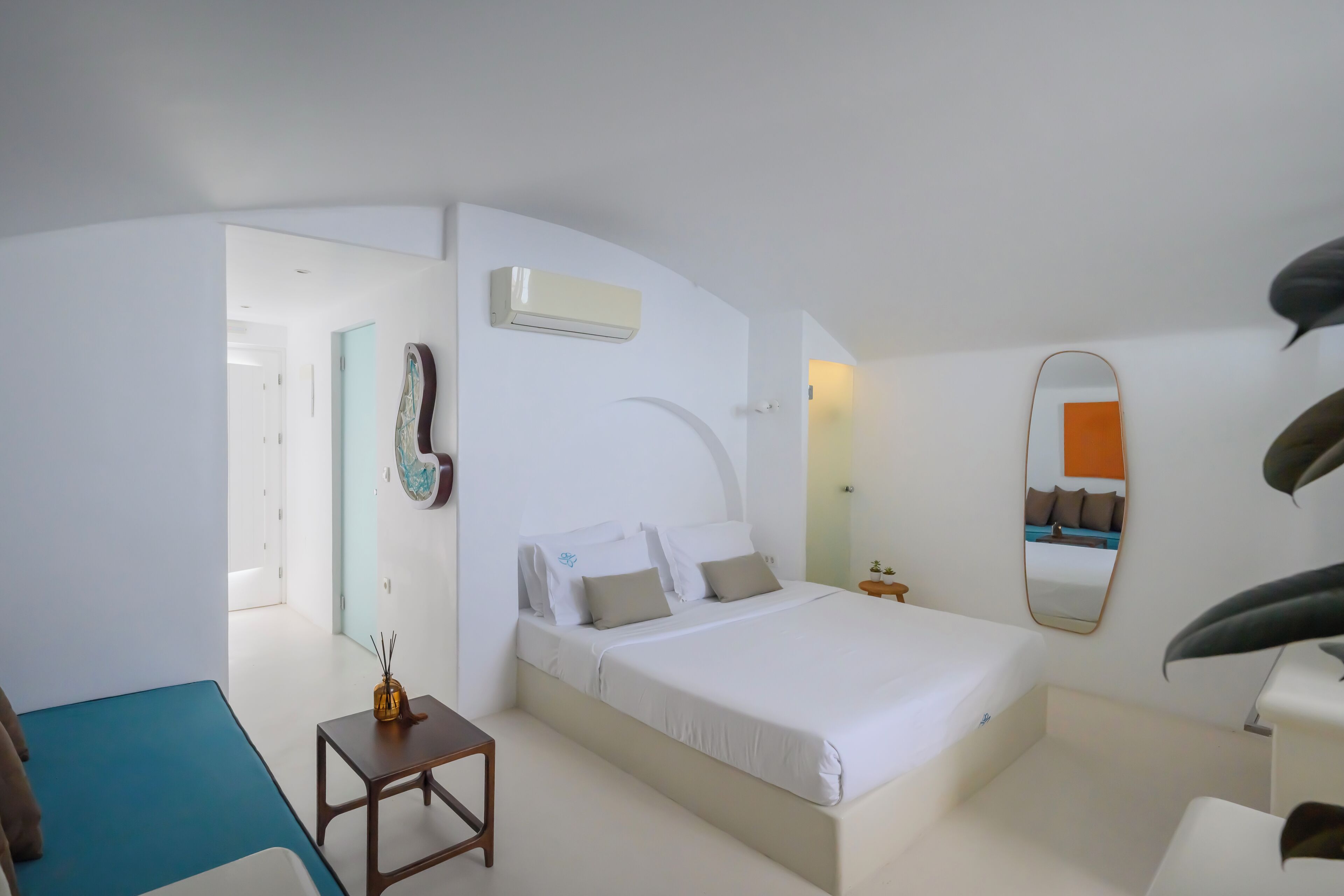 A modern hotel room with a bed, a small table, a chair, a mirror, and an air conditioning unit.