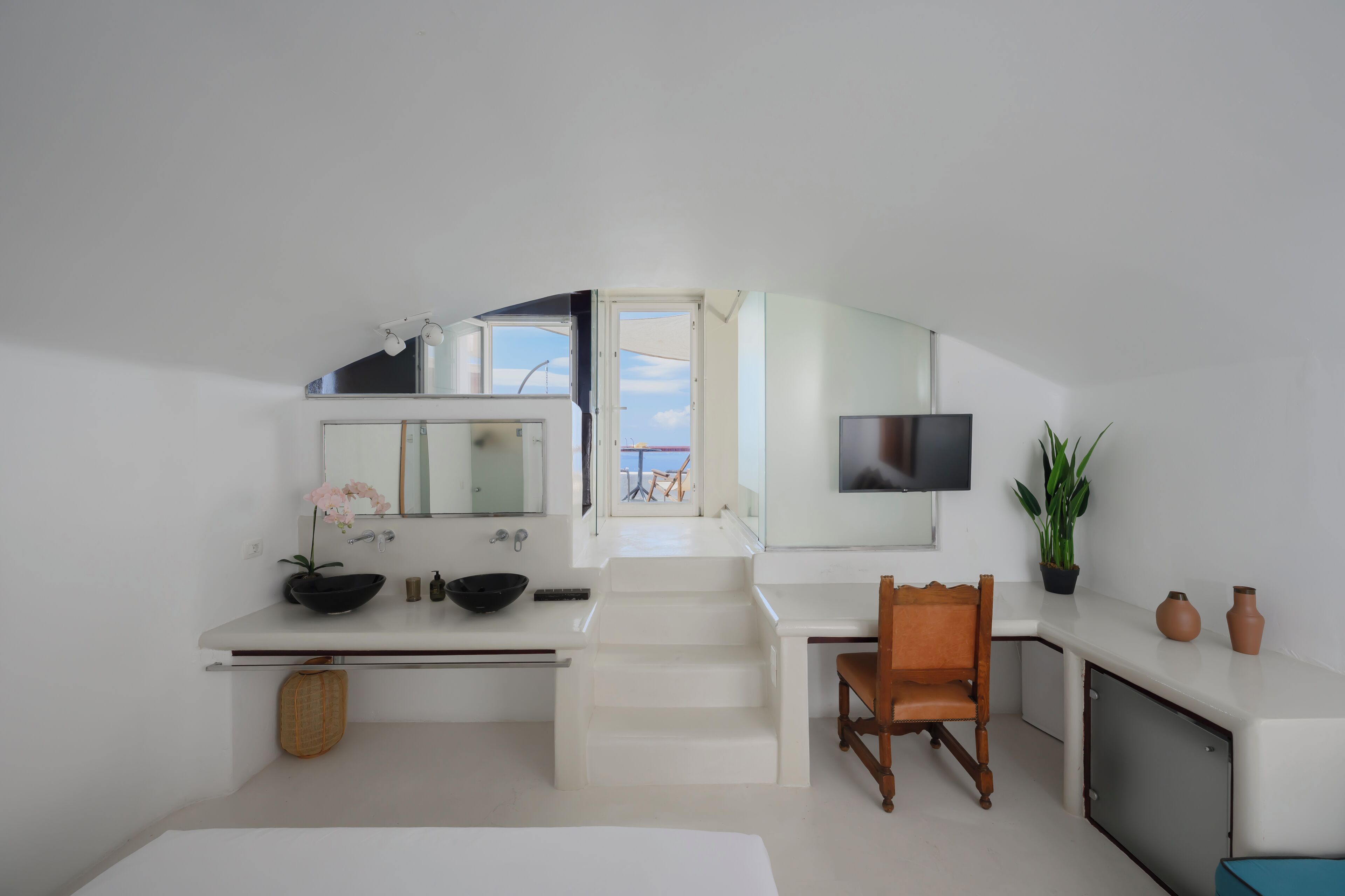 A modern hotel room with a white color scheme, a large mirror, a flat-screen TV, a wooden chair, and a view of the sea through an open door.