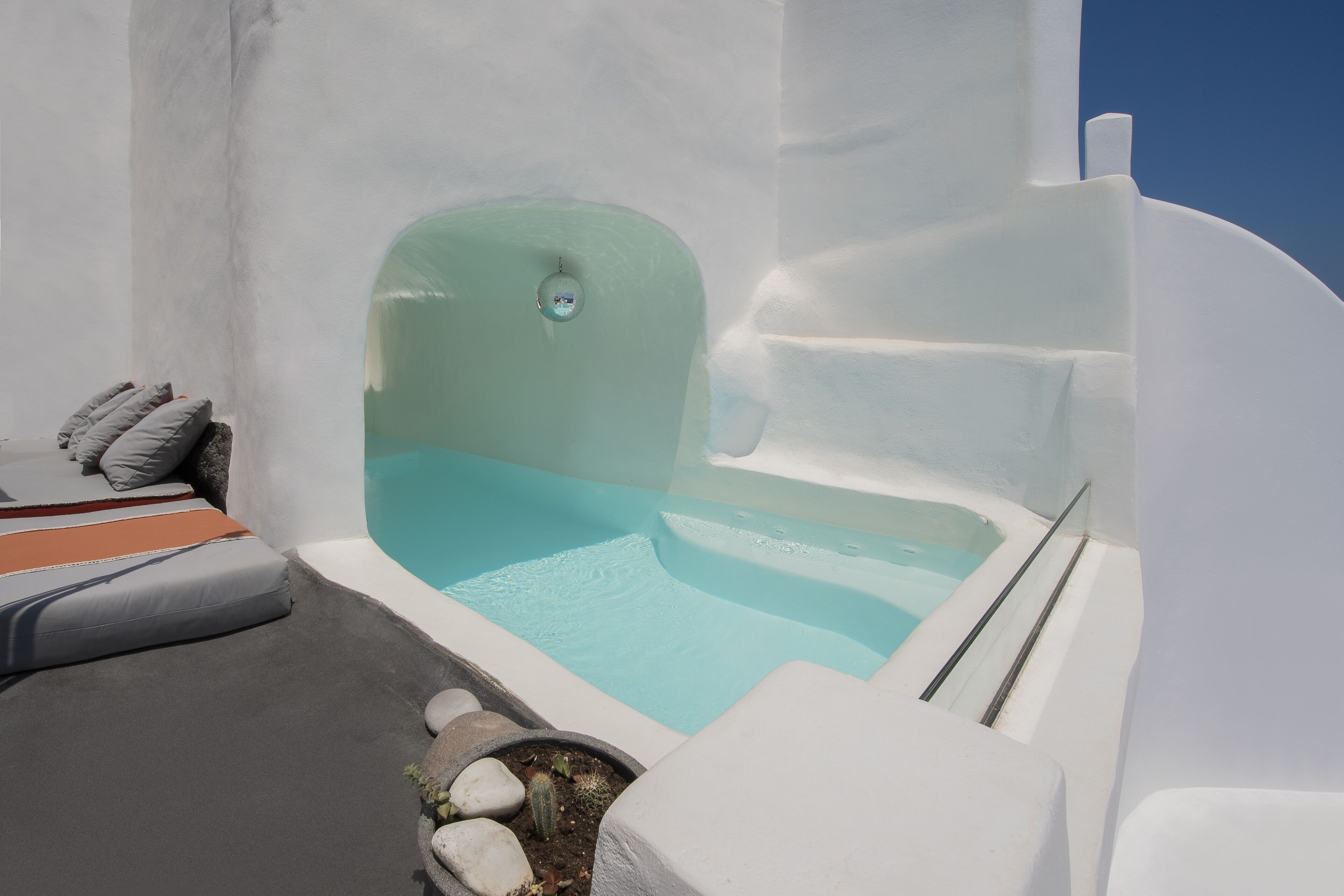Une piscine à débordement blanche, à l’eau d’un bleu cristallin, entourée de murs blancs et d’une bordure en pierre agrémentée de sièges.