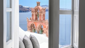 A bed with pillows, a white door, and a view of a pink building with arches and a bell tower. - Sophia Oia View (Santorini)