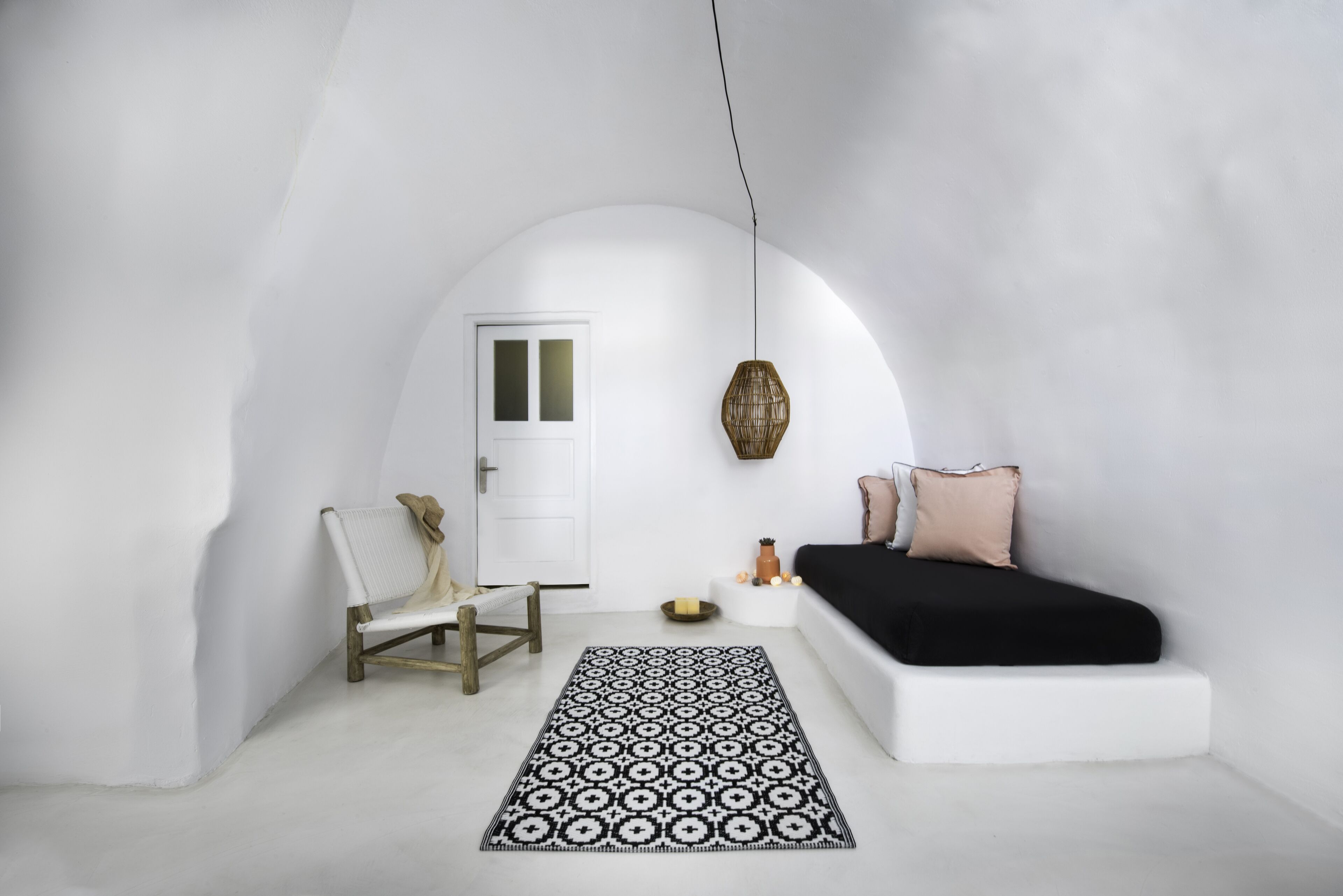Ein schlicht eingerichteter Raum mit einem weißen, bogenförmigen Durchgang, einem Holzstuhl, einem gemusterten Teppich, einem Bett mit schwarz-weißer Bettwäsche und einer Pendelleuchte.