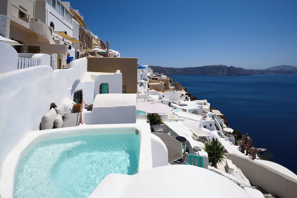 Edificio blanco con piscina, con vistas a una zona costera de aguas azules cristalinas y colinas lejanas.