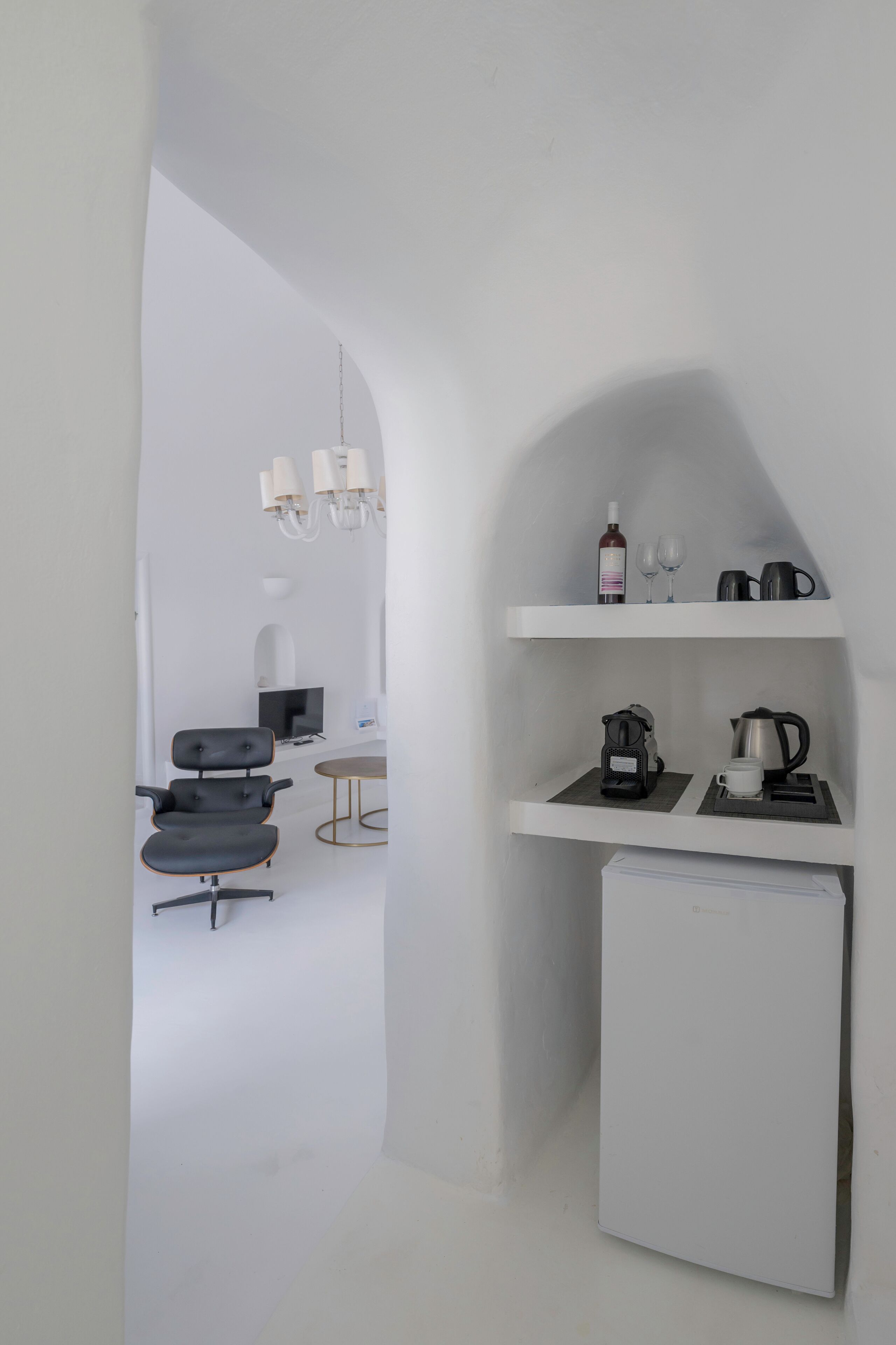 Eine kompakte Küche mit Kühlschrank, Kaffeemaschine und Wasserkocher auf der Arbeitsfläche. Im angrenzenden Sitzbereich befindet sich ein Stuhl und ein kleiner Tisch.