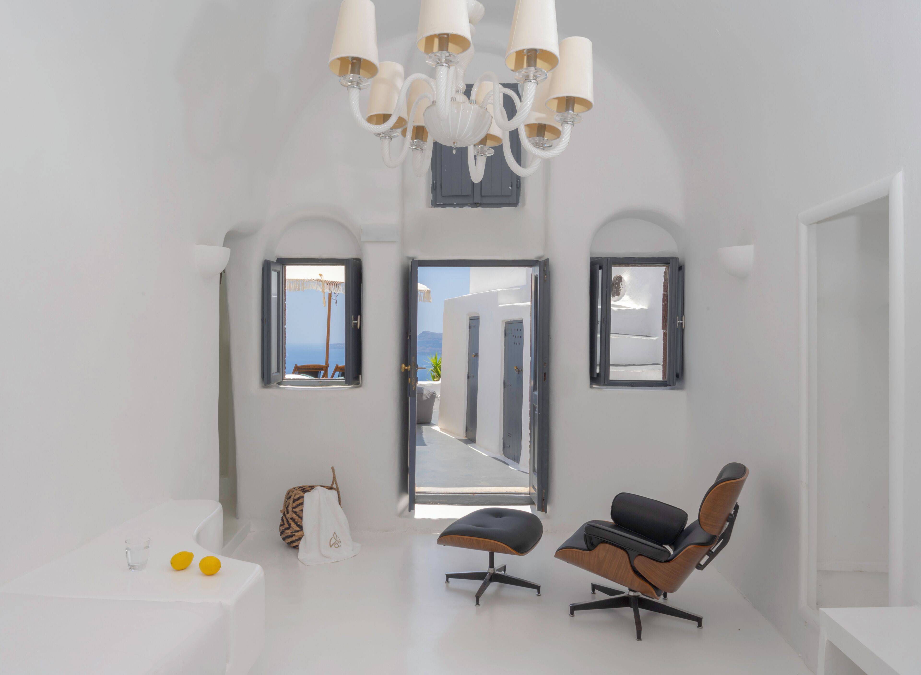 A bright, white room with a modern chandelier, two chairs, a small table, and a view of the sea through an open door.