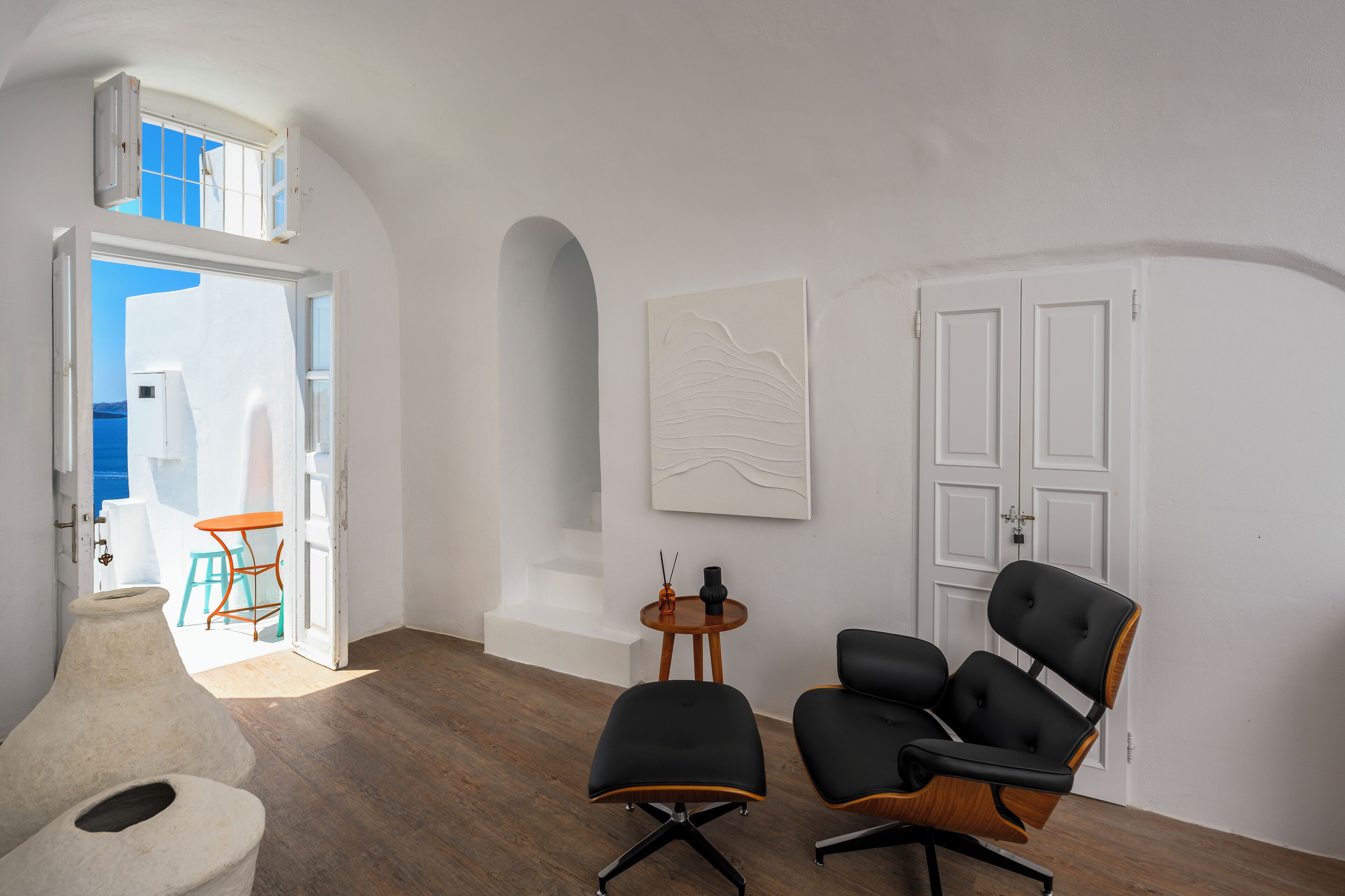 A modern interior with a wooden floor, a black leather chair, a white door, and a view of the sea through an open door.