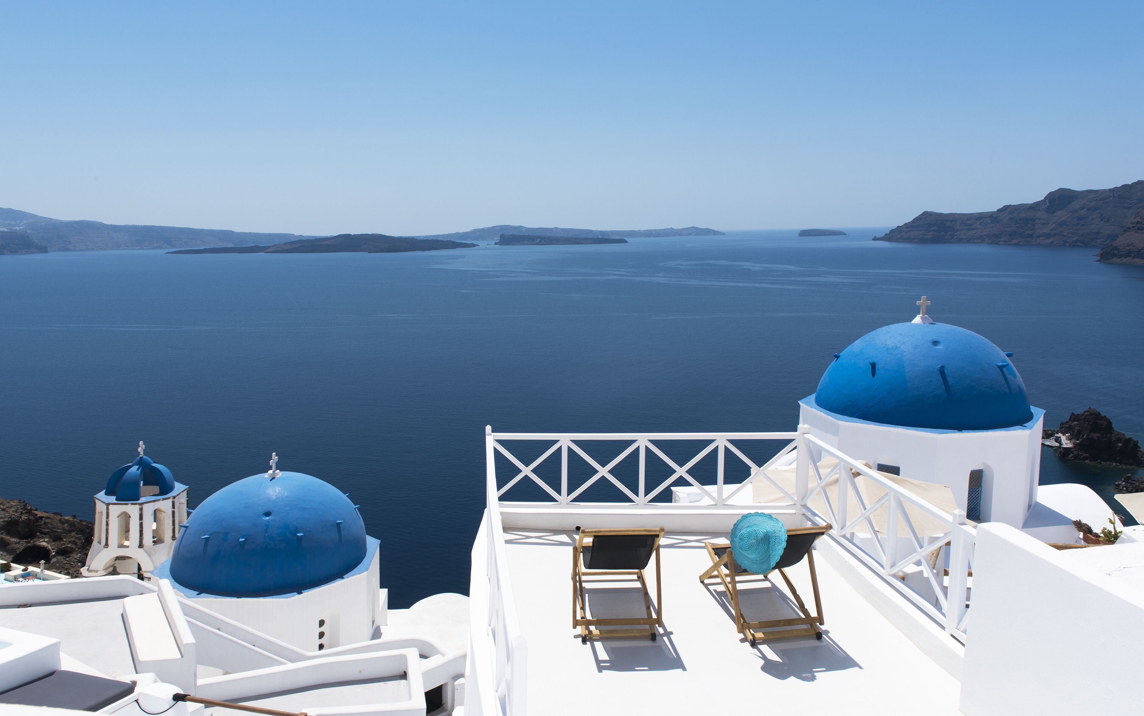 A white building with blue domes, a balcony with a chair, and a view of the sea.