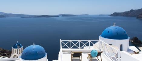 A white building with blue domes, a balcony with a chair, and a view of the sea.