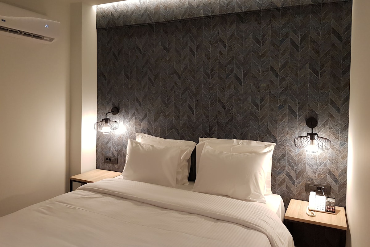 A hotel room with a bed, two bedside tables, a wall-mounted phone, and a wall-mounted air conditioner.
