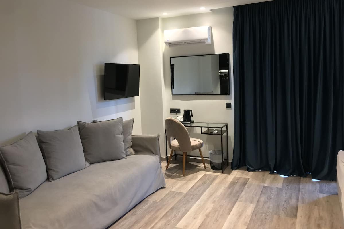 A modern hotel room with a grey sofa, a small desk with a chair, a flat-screen TV mounted on the wall, a large mirror, and a black curtain.