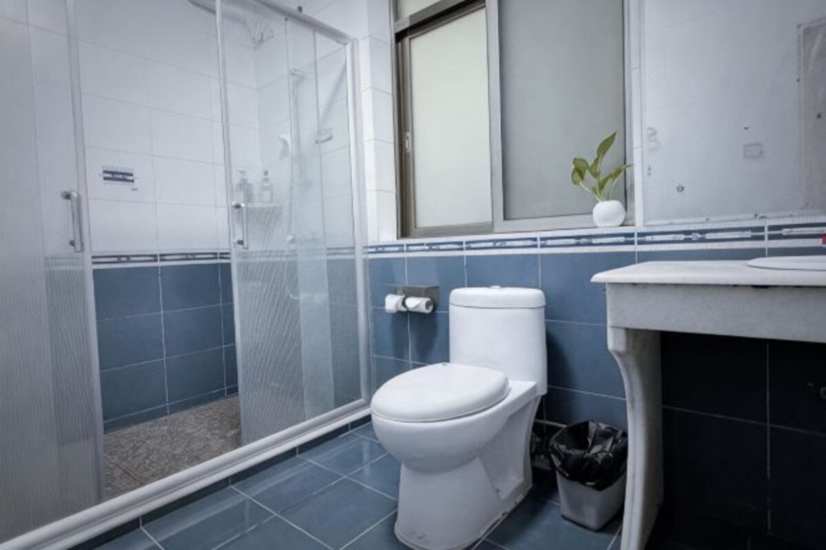 Un baño con cabina de ducha, inodoro, cubo de basura y lavamanos.