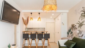 A modern living room with a flat-screen TV, a green sofa, a woven light fixture, and a kitchen area with a dining table and chairs.