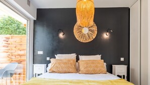 A bedroom with a large bed, two bedside tables, a wall-mounted lamp, and a window with a view of a wooden fence and greenery.