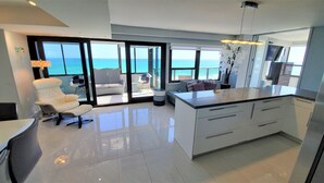 A modern kitchen with a large island, a dining area with a white chair, and a view of the ocean through large windows. - Magnificent Modern Direct Oceanfront 3 Bedroom - 1401 (Miami Beach)