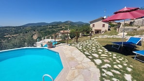 Un espace piscine doté d’un sol en pierre, offrant une vue sur les collines environnantes, et un bâtiment surmonté d’un parasol rouge.