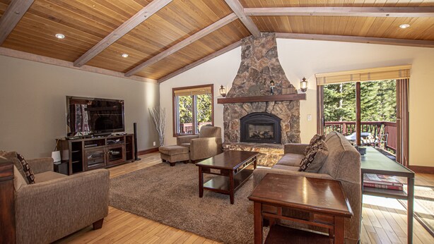 A spacious living room with a stone fireplace, wooden ceiling beams, and a large window overlooking a deck. - Highlands Lodge by Hauserman Rental Group (Tahoe City)