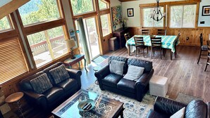 Uma sala de estar ampla com teto alto, grandes janelas e uma área de jantar com mesa e cadeiras.