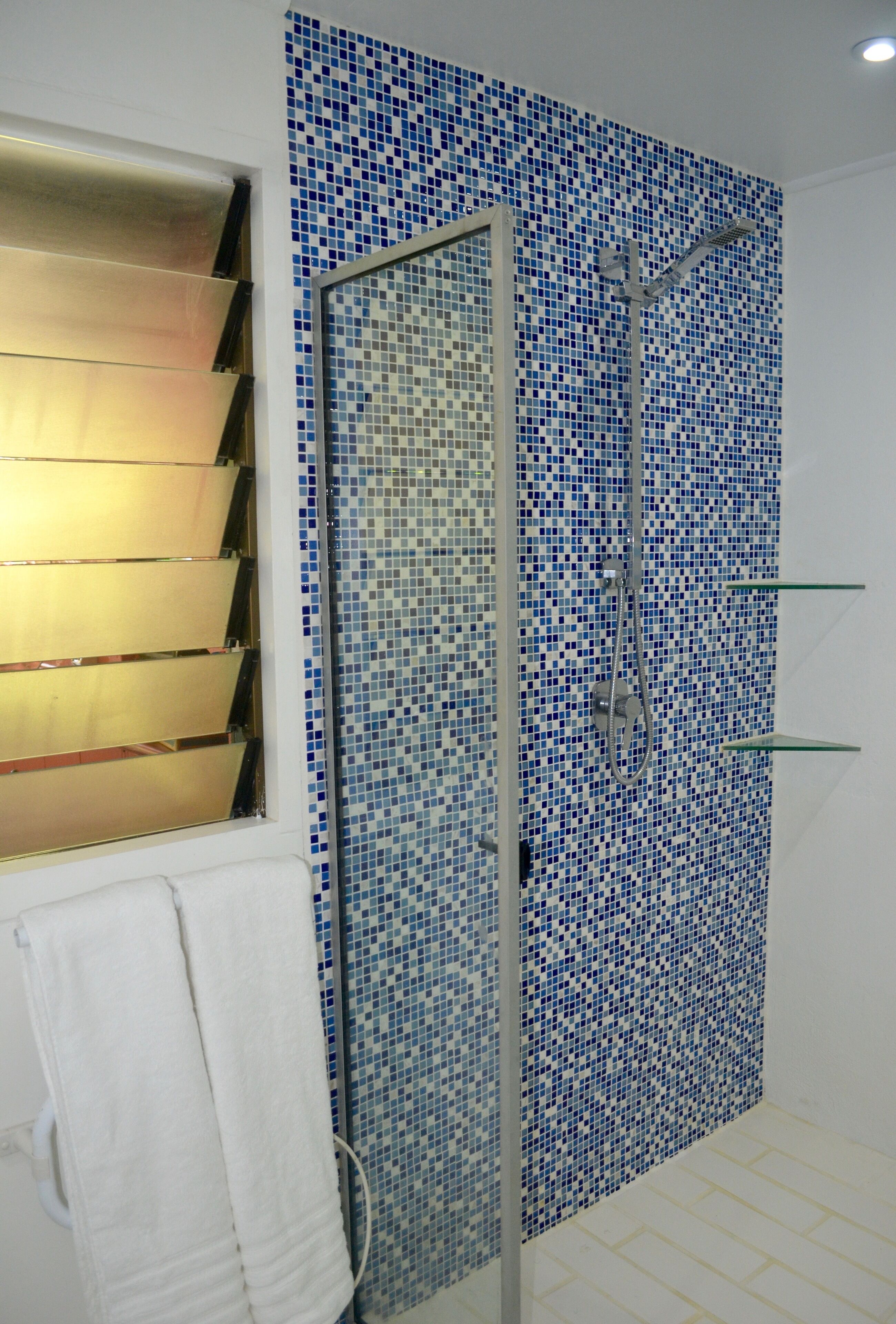 A bathroom with a shower area featuring blue mosaic tiles, a glass shower door, and a towel rack with a white towel.