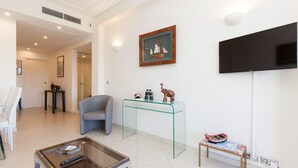 A modern living room with a glass coffee table, a blue armchair, a glass console table with decorative items, and a flat-screen TV mounted on the wall. - Sea View, Cannes Croisette, in Front of the Sea (Cannes)