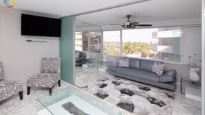 A modern living room with a grey sofa, patterned armchairs, a glass coffee table, and a large window offering a city view.