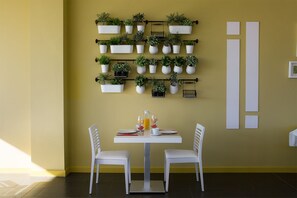 Un coin repas avec une table et des chaises blanches, un présentoir à plantes fixé au mur et une table préparée pour le petit-déjeuner.