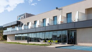 A modern hotel with a flat facade, large glass windows, outdoor seating, and a parking area. - NRuta Ponferrada (Bembibre)