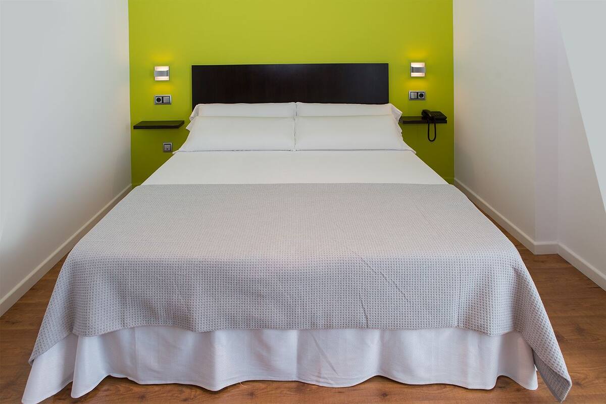 A neatly made bed with a white duvet and a grey textured cover, set against a green wall with two bedside lamps and a phone on a nightstand.