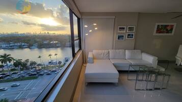 A modern living room with a white sofa, a glass coffee table, and a view of a waterfront with boats and cars.