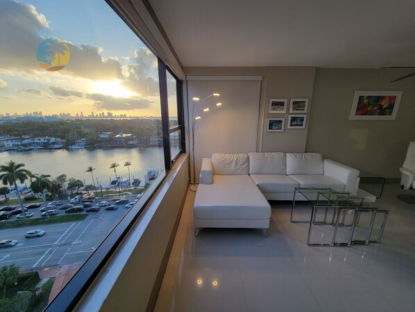 A modern living room with a white sofa, a glass coffee table, and a view of a waterfront with boats and cars.
